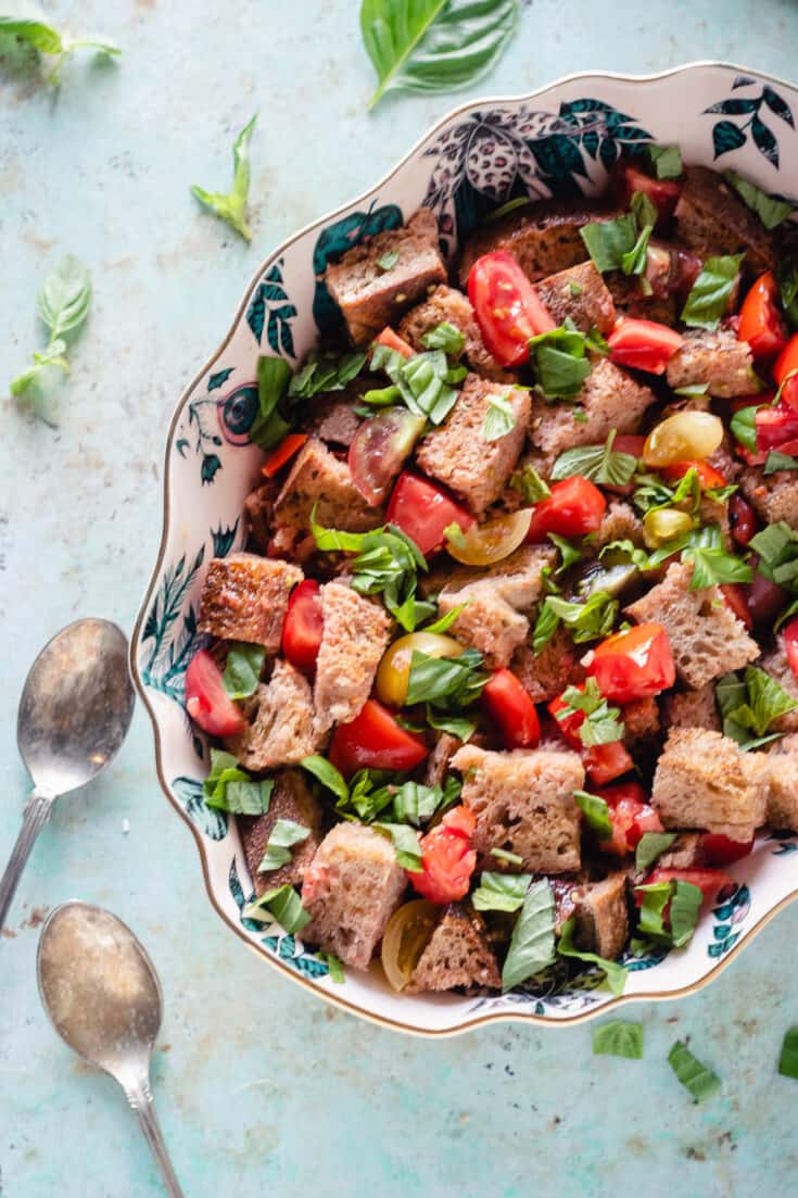 Panzanella in a patterned oval bowl with a pair of serving spoons on the counter beside it