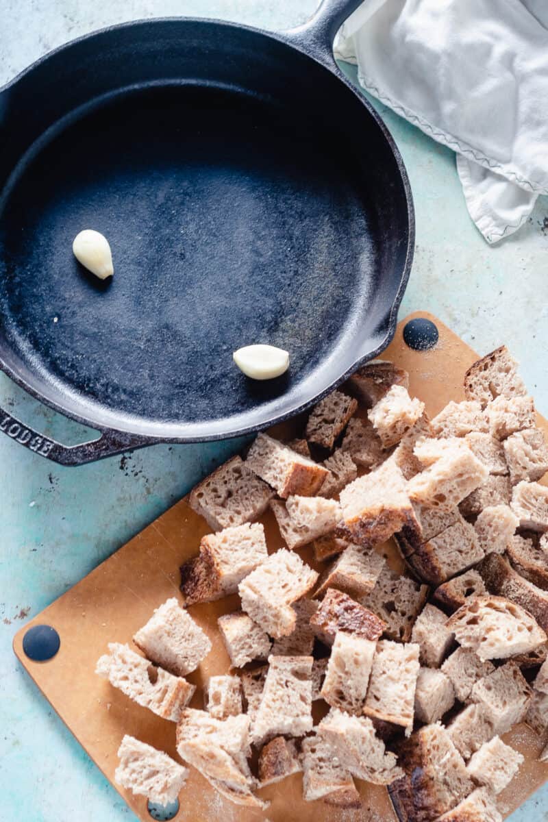 Garlic cloves in a cast iron skillet and cubes of bread on a cutting board next to it