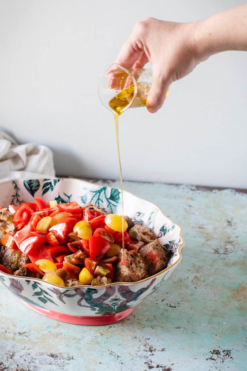 Hand pouring olive oil over panzanella in a patterned oval bowl