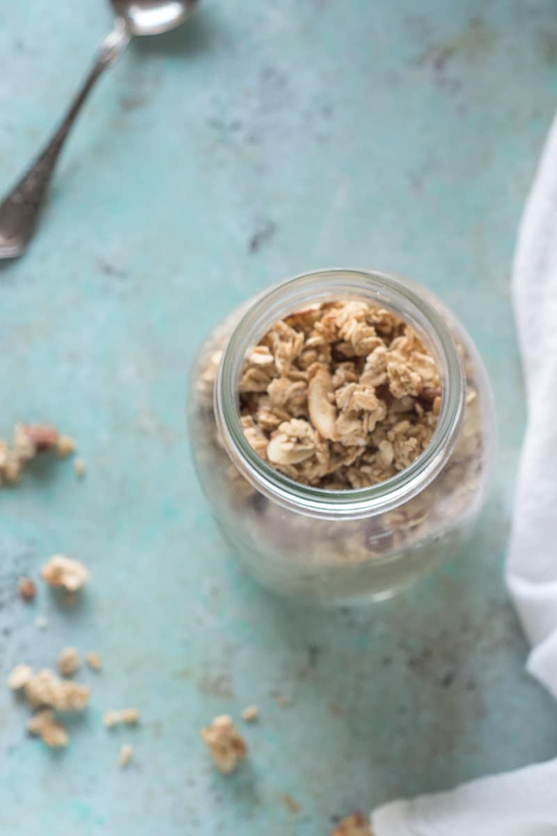 Maple Nut Granola in a jar, with a spoon and tea towel in the background, overhead shot