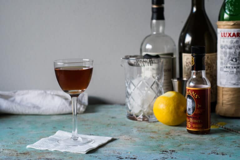 Martinez Cocktail in a glass with a mixing glass, lemon, bitters and bottles behind it