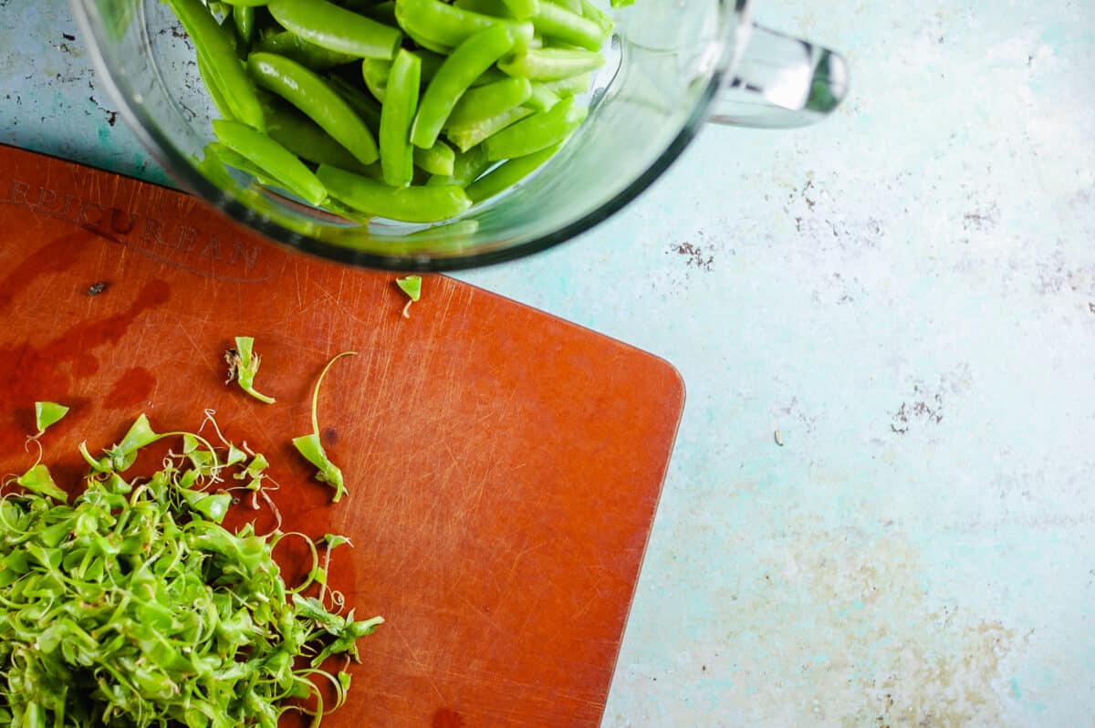 Sugar snap pea trimmings on a cutting board with a bowl of peas next to it