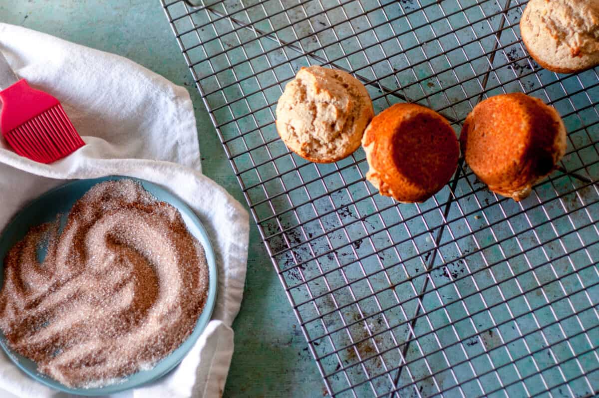 Muffins on a cooling rack with a cinnamon sugar mixture in a blue dish