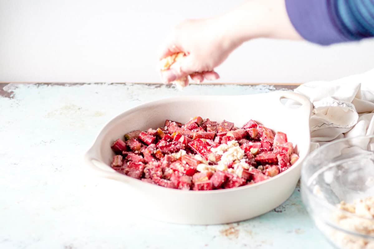 Hand adding crumble topping to a rhubarb in a baking dish
