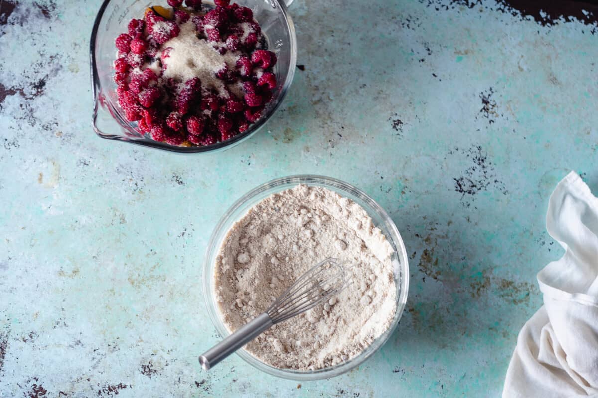 Raspberries with sugar in a mixing bowl and flour in a separate bowl with a whisk