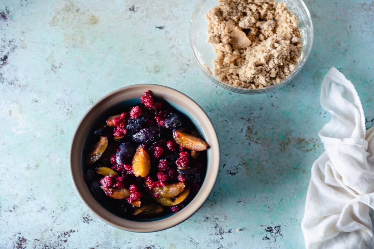 Raspberries and plums tossed with sugar in a baking dish with crisp topping in a bowl beside it