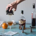 Revolver Cocktail being strained into a glass with jiggers, an orange, and whiskey and coffee liqueur in the background