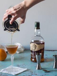 Revolver Cocktail being strained into a glass with jiggers, an orange, and whiskey and coffee liqueur in the background