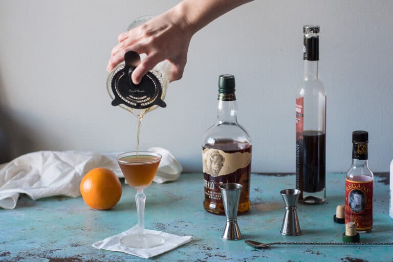 Revolver Cocktail being strained into a glass with jiggers, an orange, and whiskey and coffee liqueur in the background