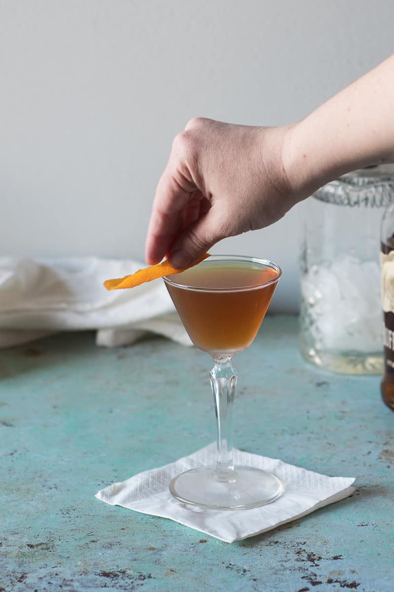 Hand expressing orange peel over the rim of the glass of the Revolver cocktail