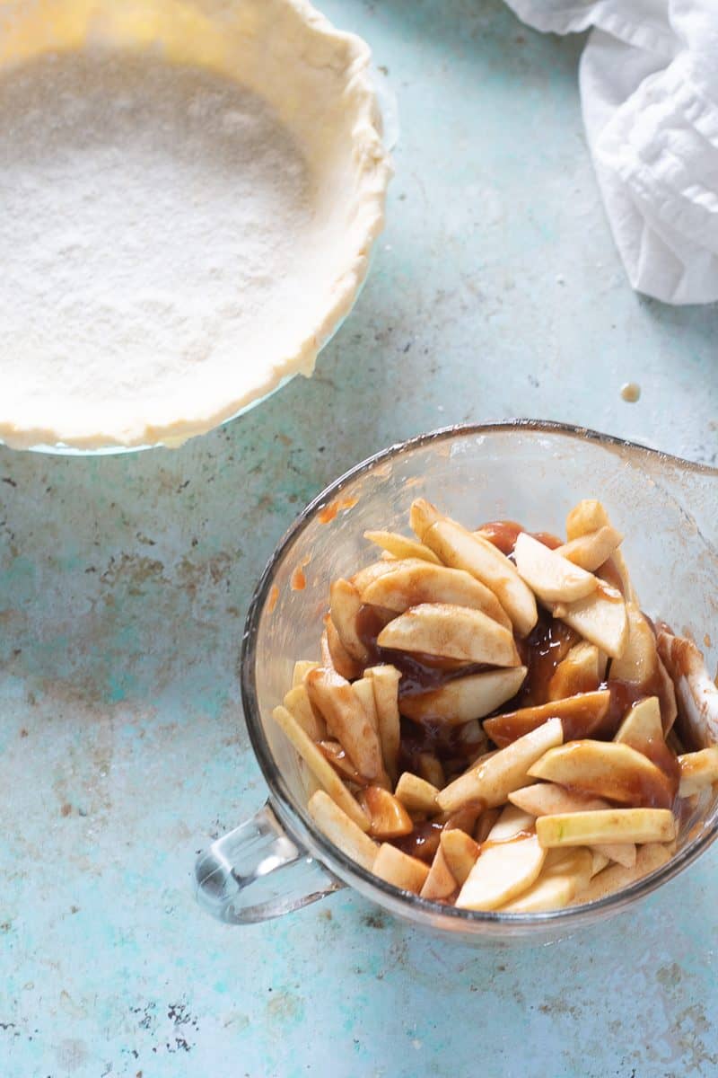 Unbaked pie shell with crust dust and apple filling in a bowl beside it