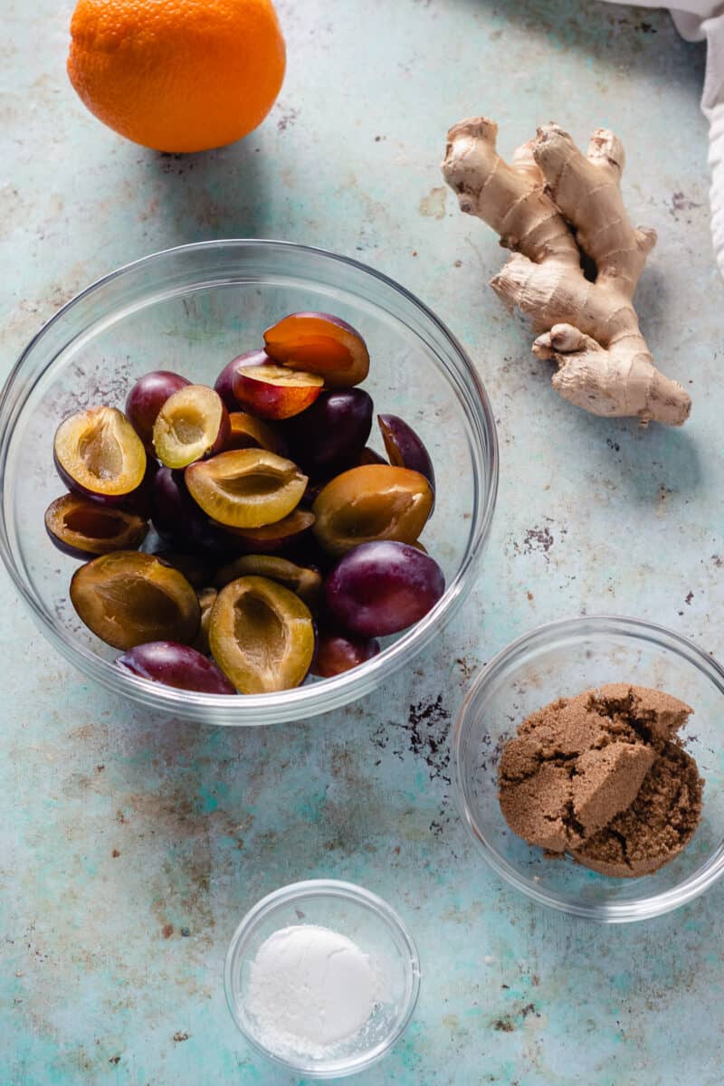 An orange, a bowl of halved plums, ginger root, brown sugar