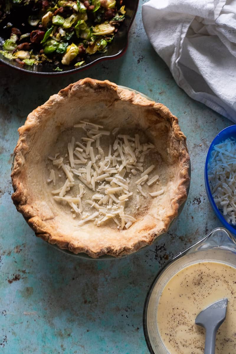 Pie crust with some shredded gruyere cheese in the bottom next to a bowl of eggy custard filling and a skillet with bacon and Brussels sprouts