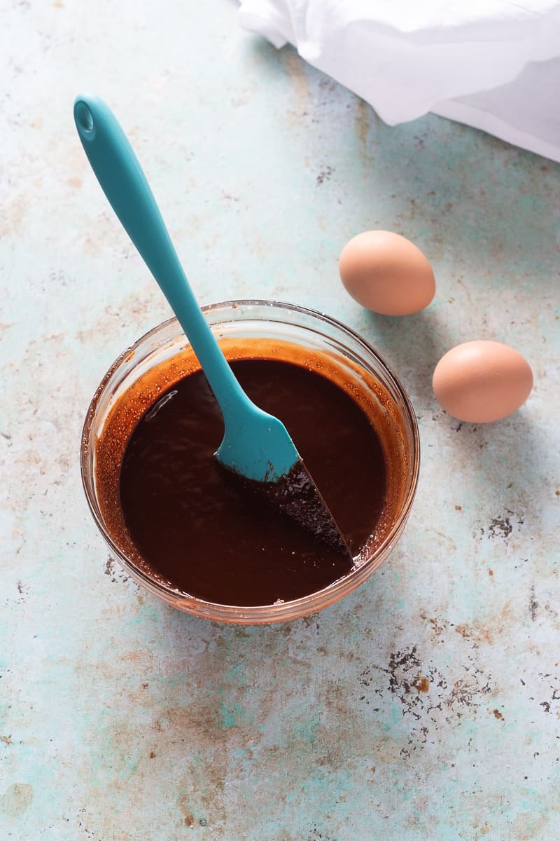 A bowl of melted chocolate, butter, and sugar with two eggs sitting near by