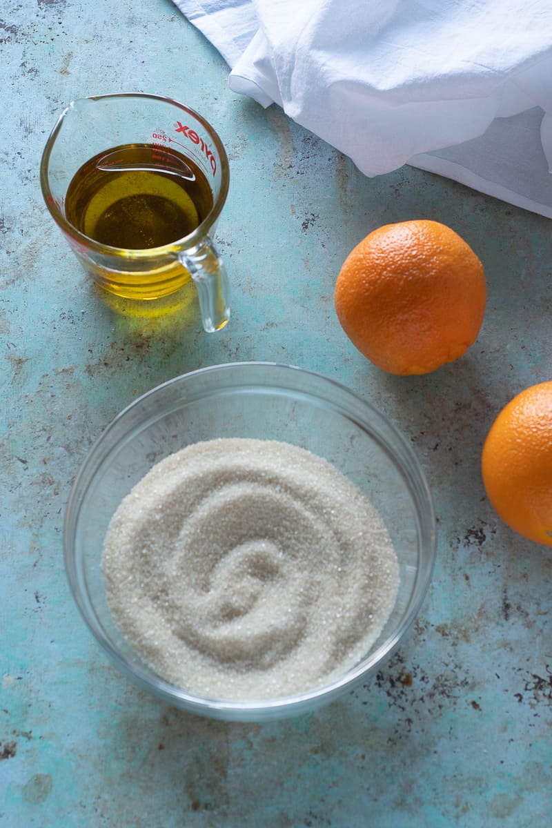 Olive oil in a measuring cup next to two navel oranges and a bowl of sugar