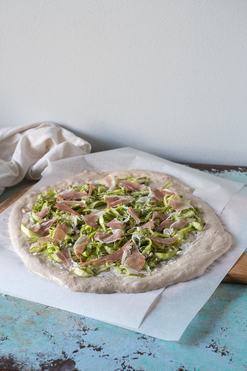 Prosciutto and shaved asparagus on unbaked pizza dough on parchment paper on a wooden pizza peel