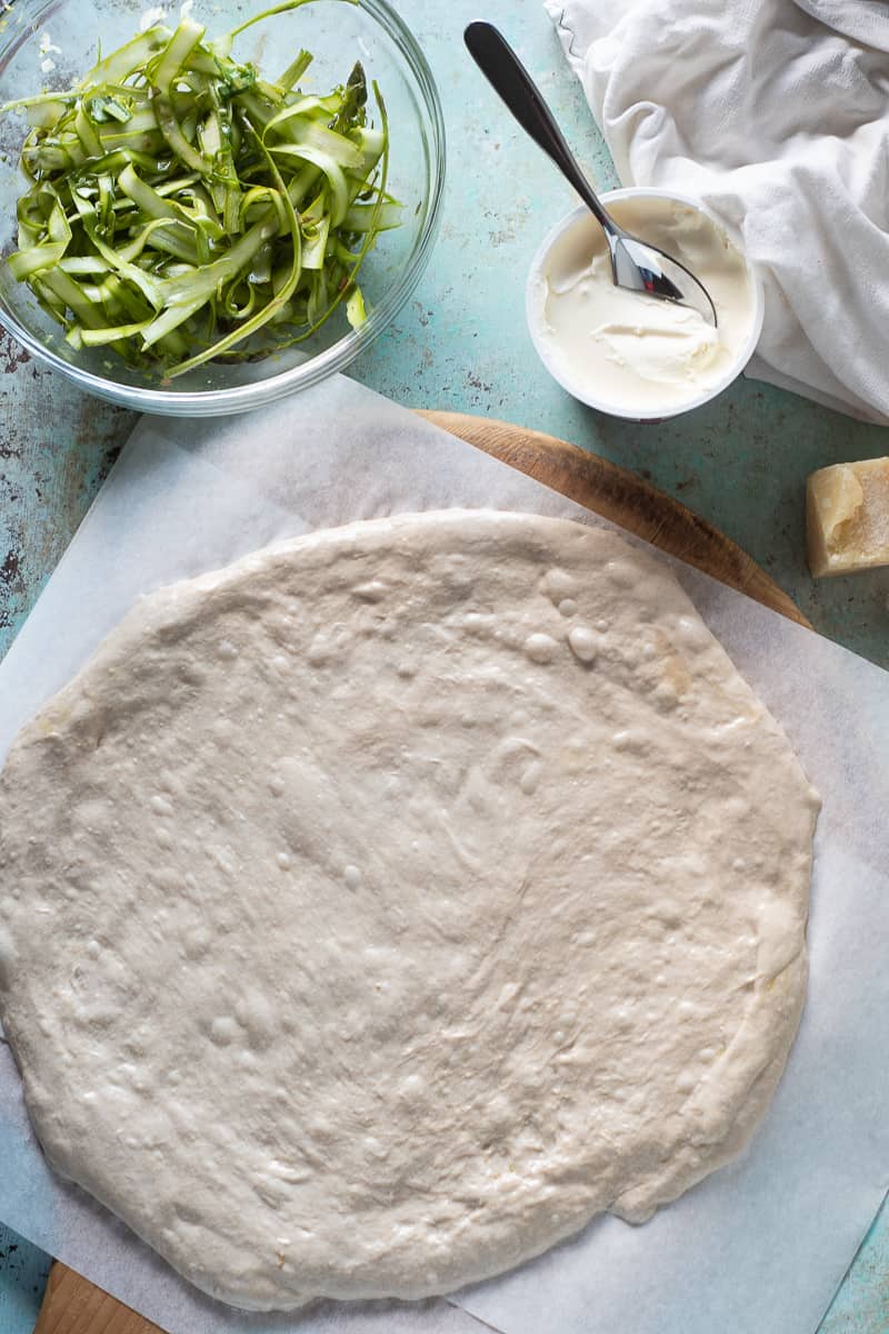Shaved asparagus in a bowl, creme fraiche, a hunk of parmesan cheese, and pizza dough stretched out on parchment paper