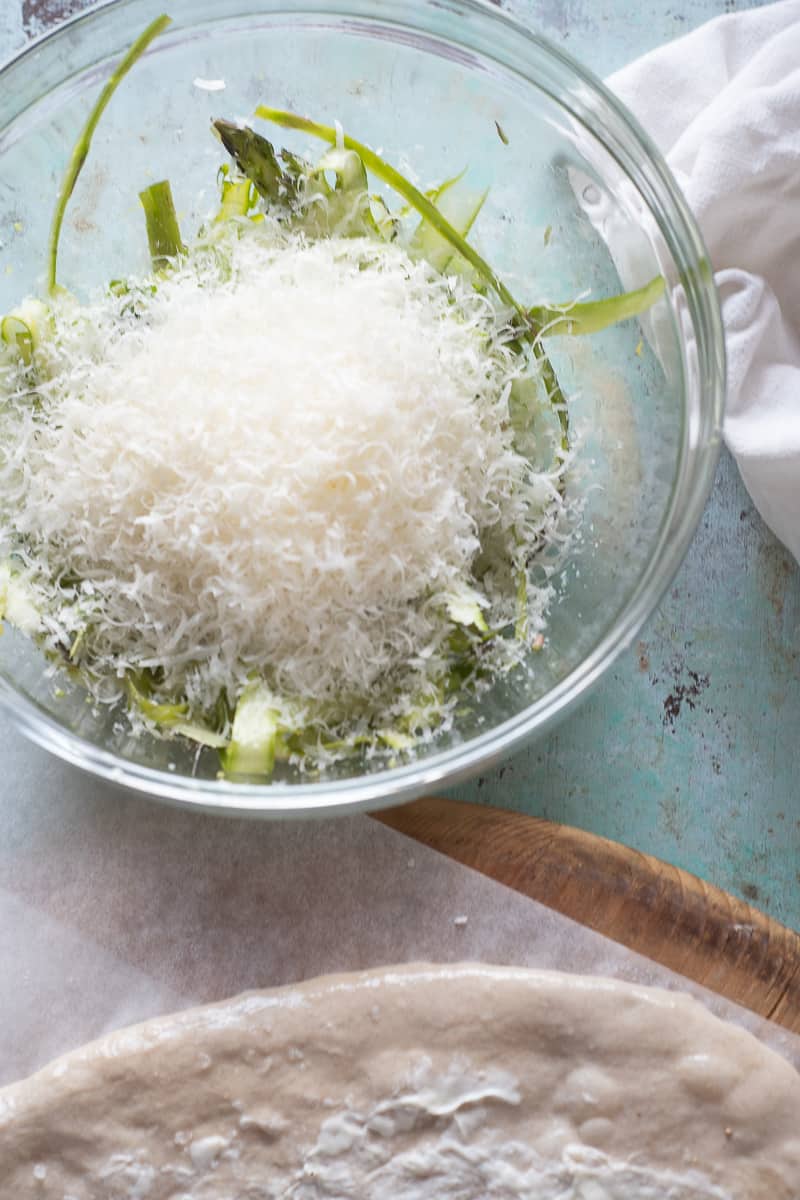 A pile of grated parmesan cheese over a bowl of shaved asparagus next to the edge of some pizza dough