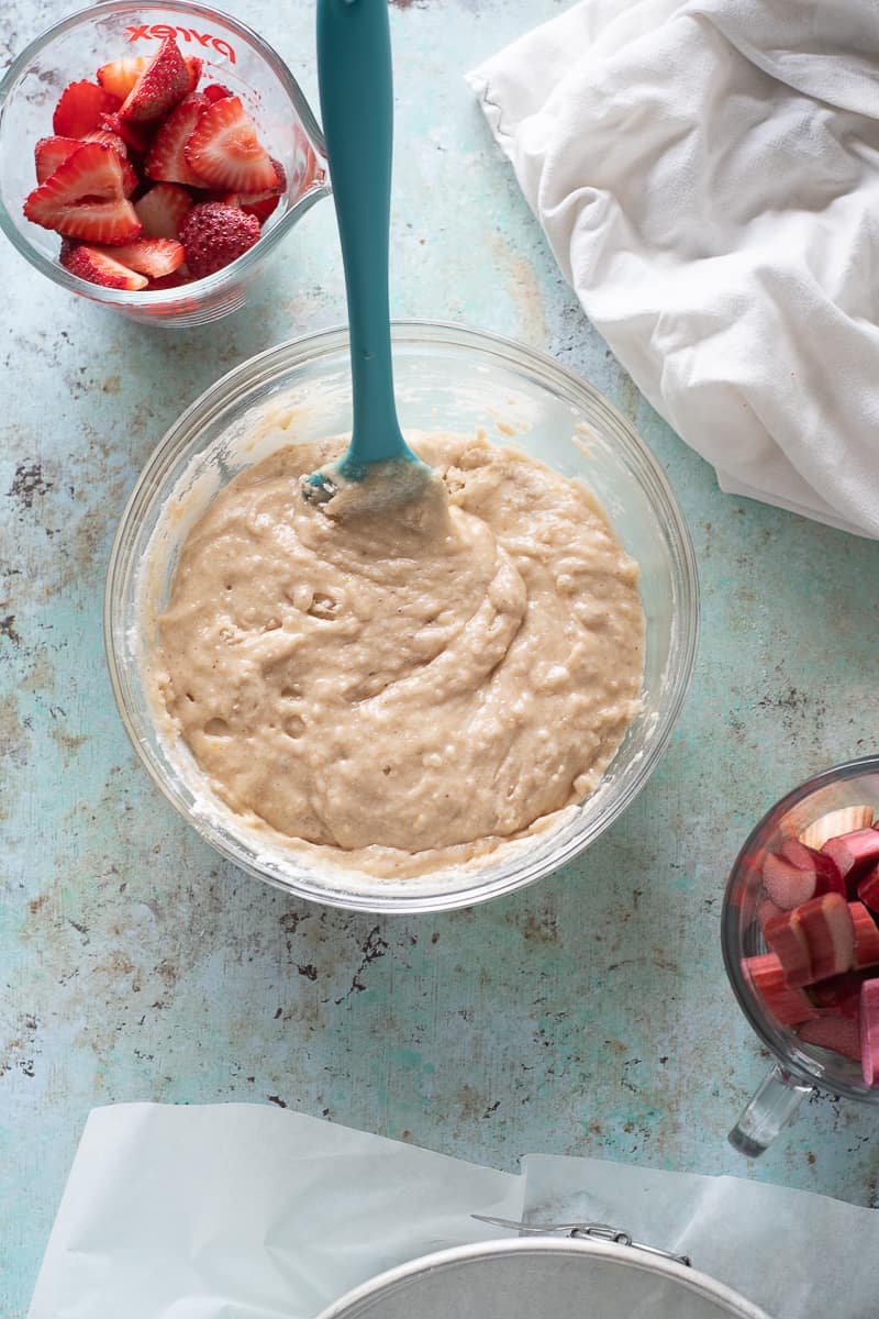 Sliced strawberries in a measuring cup next to a bowl of thick batter with sliced rhubarb in another measuring cup