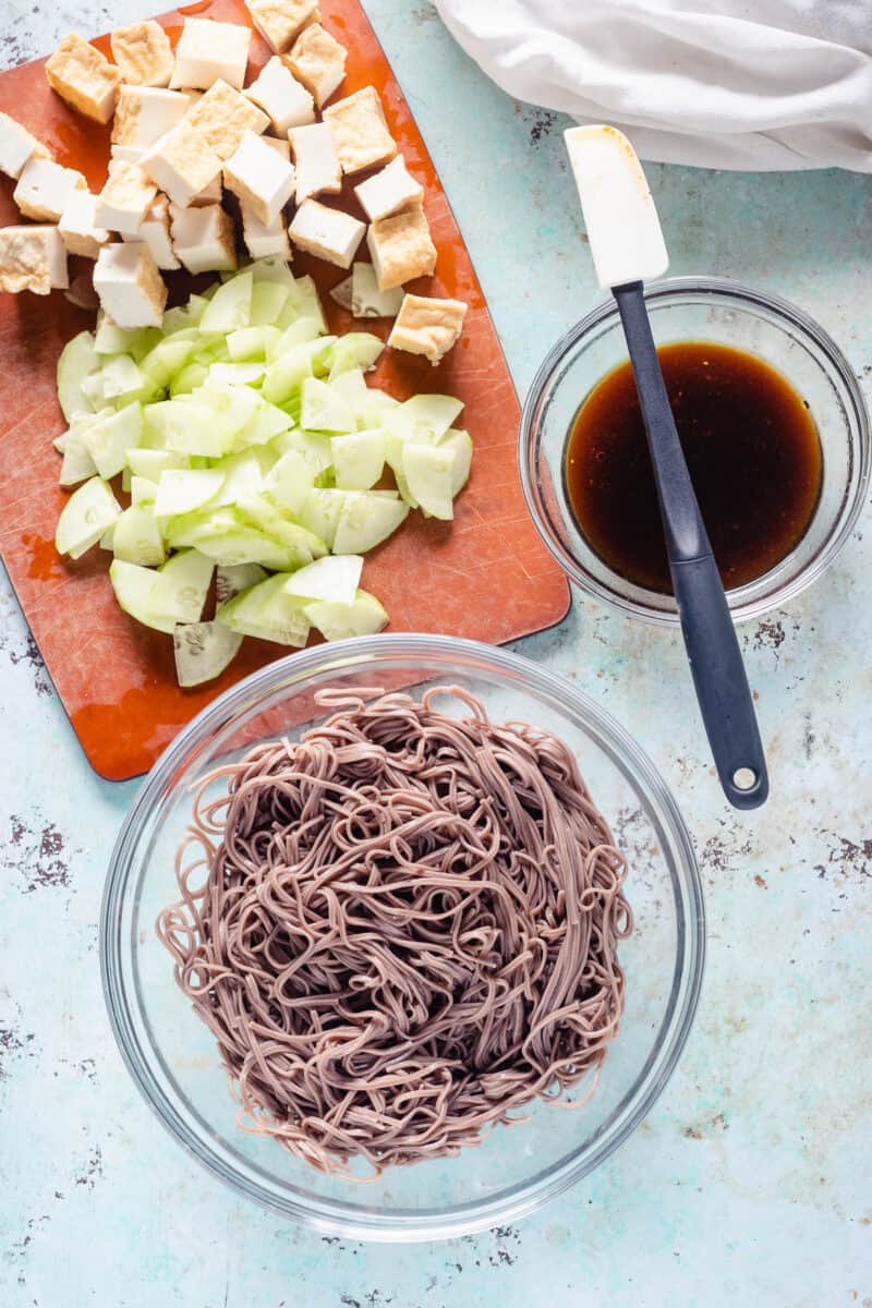 Chopped tofu, sliced cucumbers, a bowl of sesame-lime sauce, and cooked soba noodles in a bowl