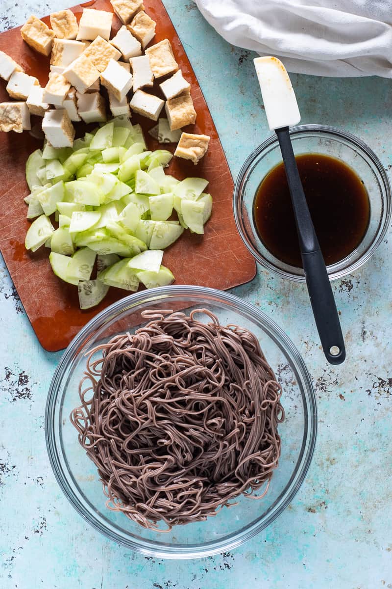 Sliced tofu and cucumbers on a cutting board, a bowl of sesame lime sauce, and a big bowl of cold soba noodles