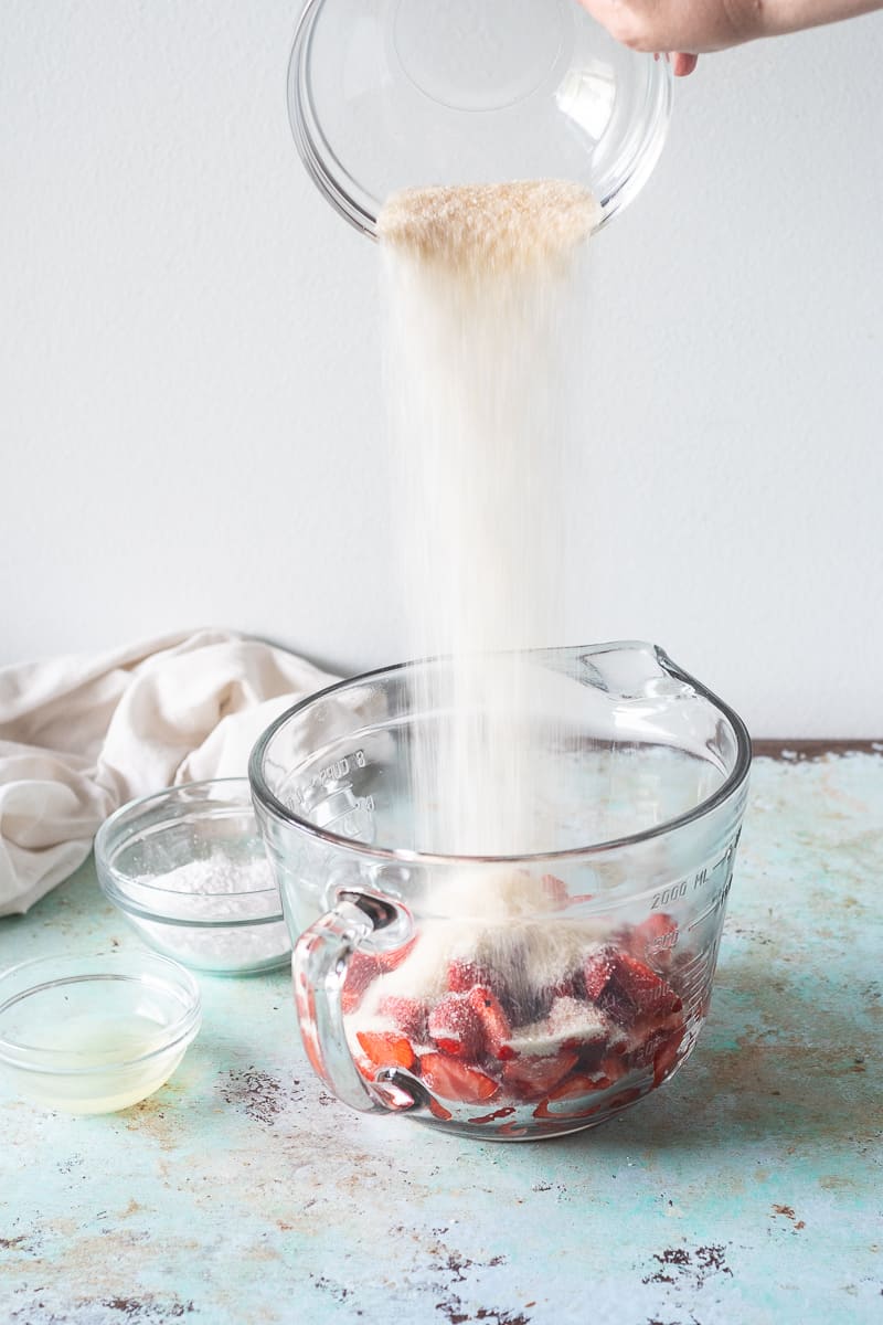 Hand pouring sugar over a bowl of sliced strawberries