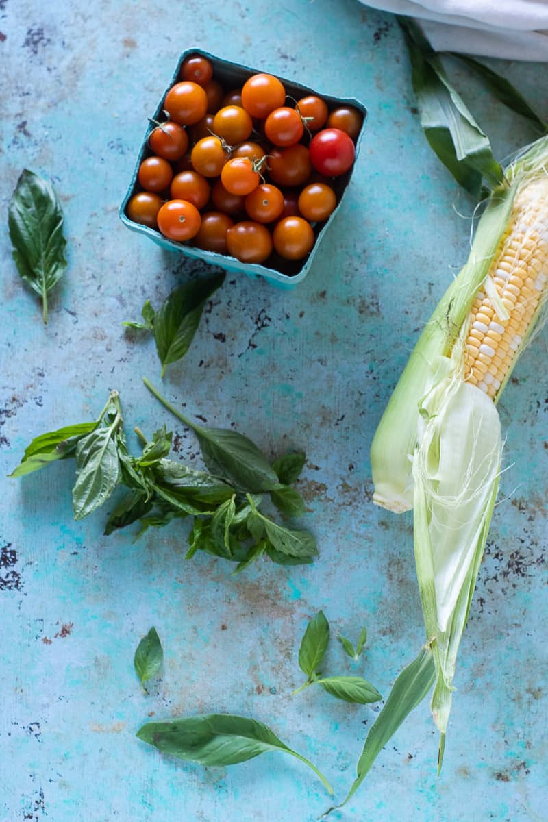 Pint of sungold tomatoes, ear of corn partially shucked, basil leaves on a table