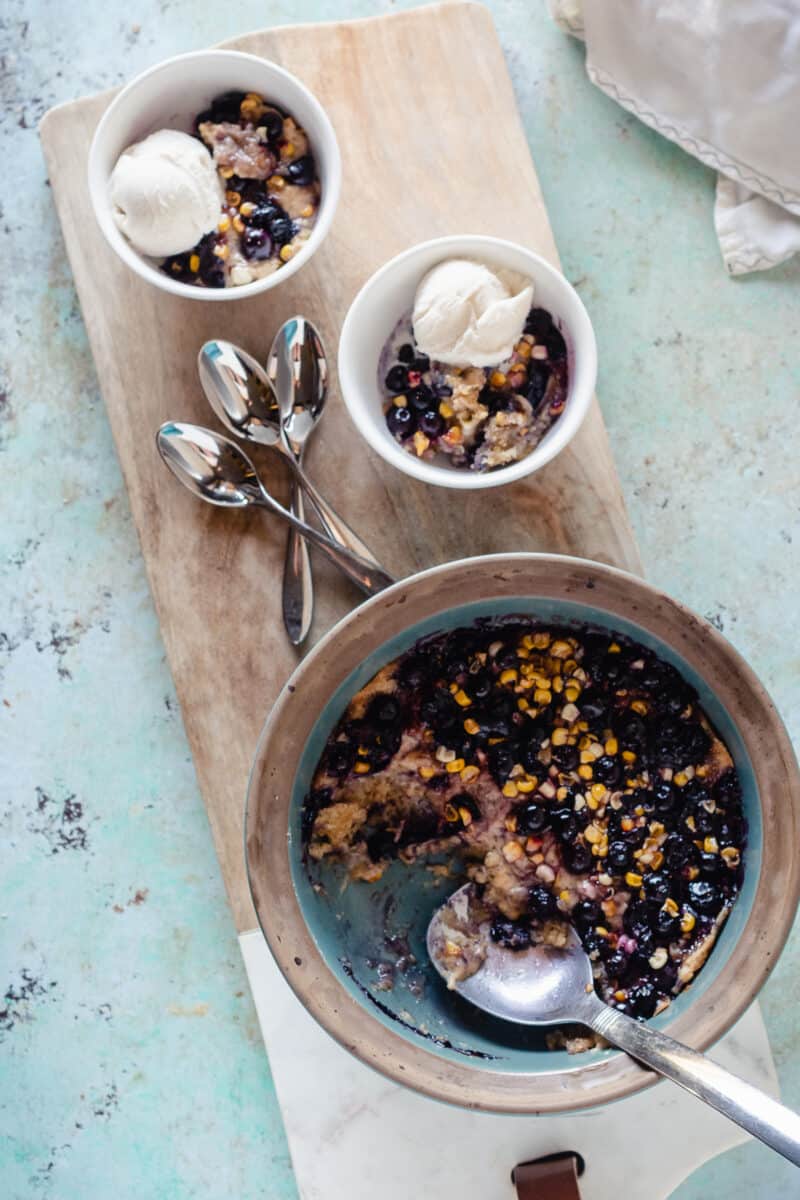 Two bowls of sweet corn & blueberry spoon cake with vanilla ice cream alongside a larger dish of spoon cake