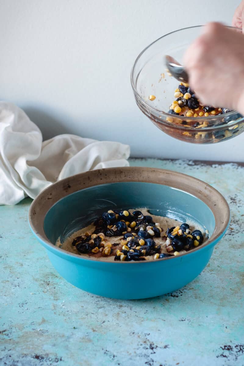 Hand spooning sweet corn and blueberry topping over the spoon cake batter