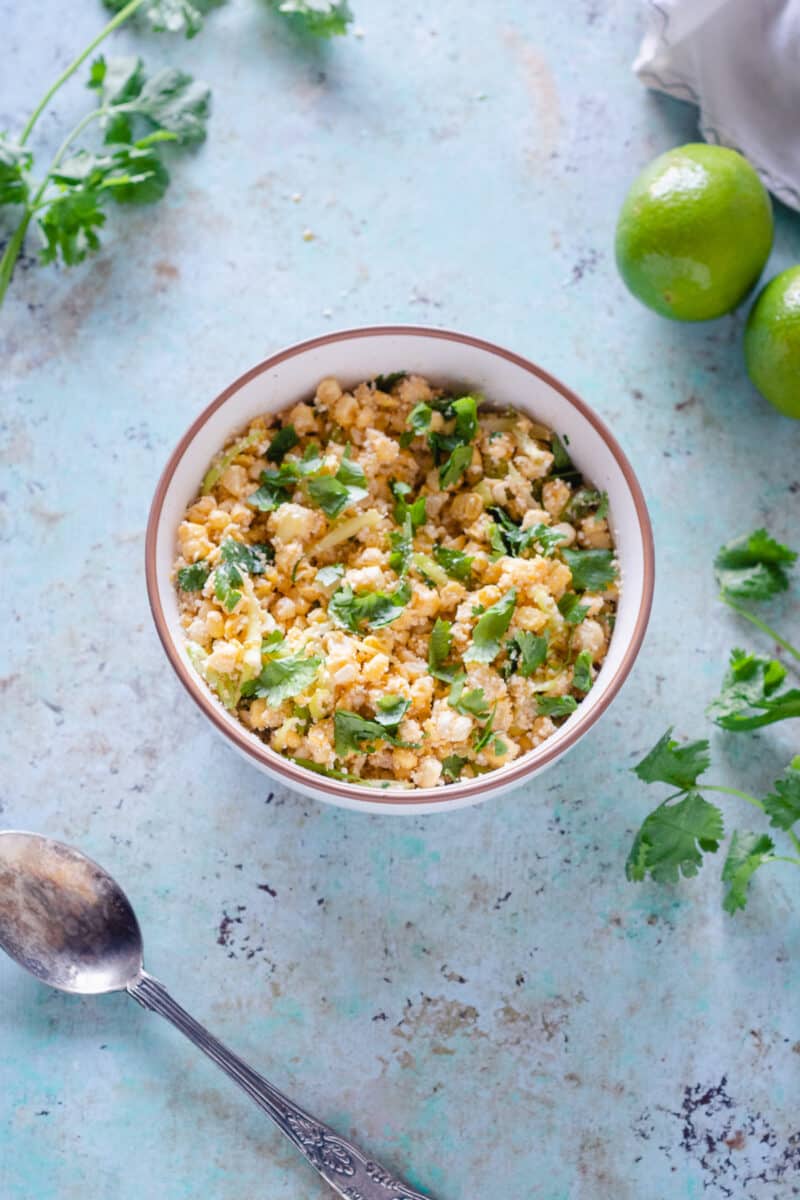 Corn with miso lime butter in a white bowl with a vintage serving spoon and cilantro leaves and a few limes scattered around, overhead view
