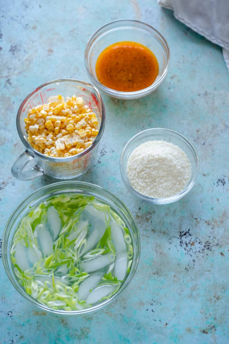 Corn kernels in a glass measuring cup, miso lime butter in a small glass bowl, grated cotija cheese in a glass bowl, thinly sliced scallions in glass bowl with ice water