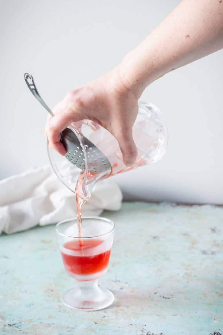 Hand straining Negroni into a footed rocks glass from a mixing glass