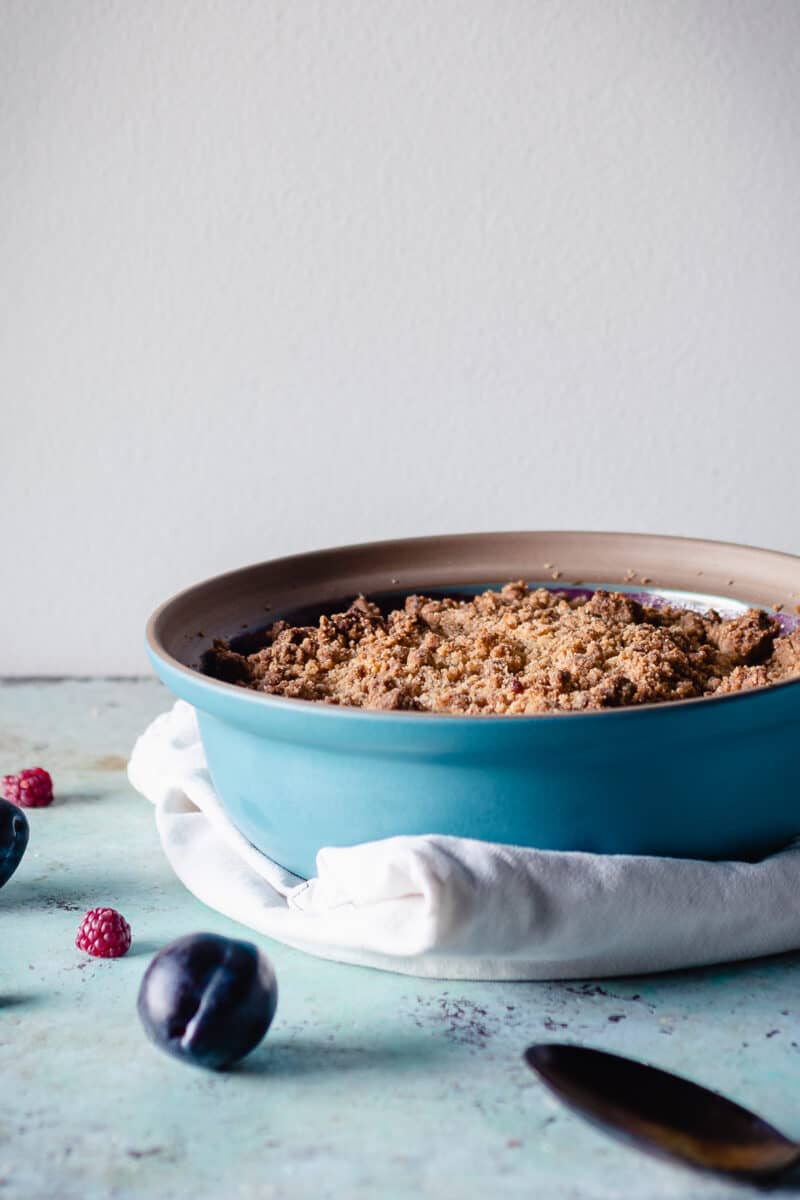 Raspberry Plum Crisp in a blue baking dish with plums and raspberries scattered nearby