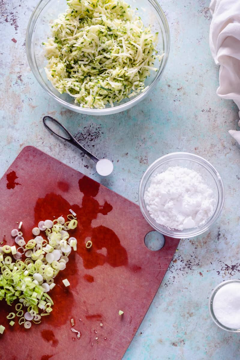 Shredded zucchini in a bowl, baking powder in a measuring spoon, potato starch in a bowl, and sliced scallions on a cutting board