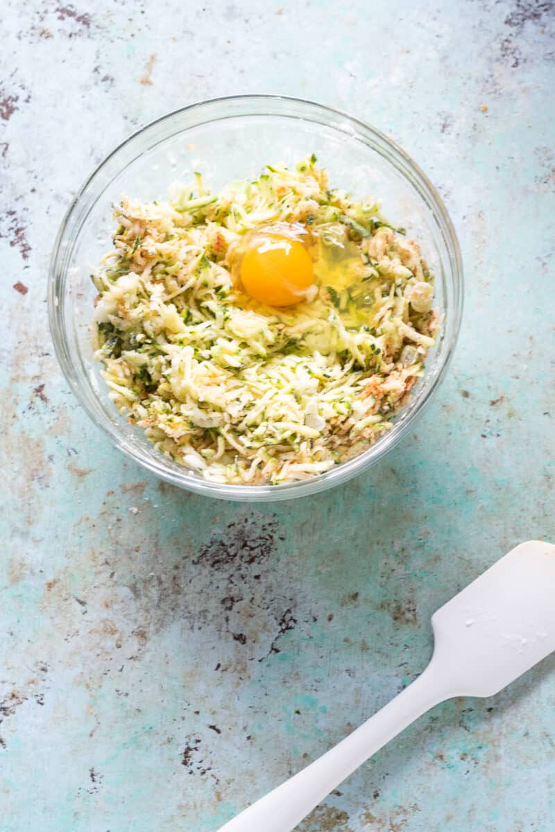 Shredded zucchini in a bowl with a mixture of potato starch, spices, and an egg on top