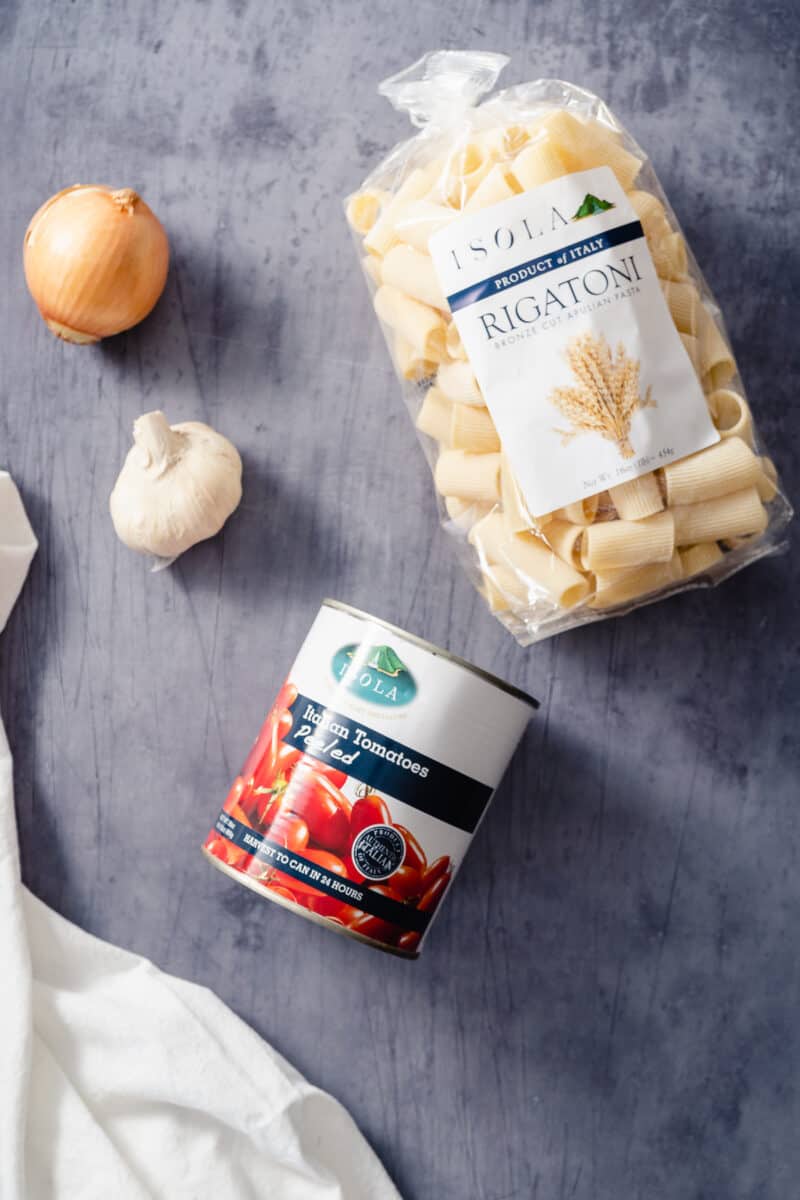 Whole peeled tomatoes, a garlic bulb, a yellow onion, and rigatoni