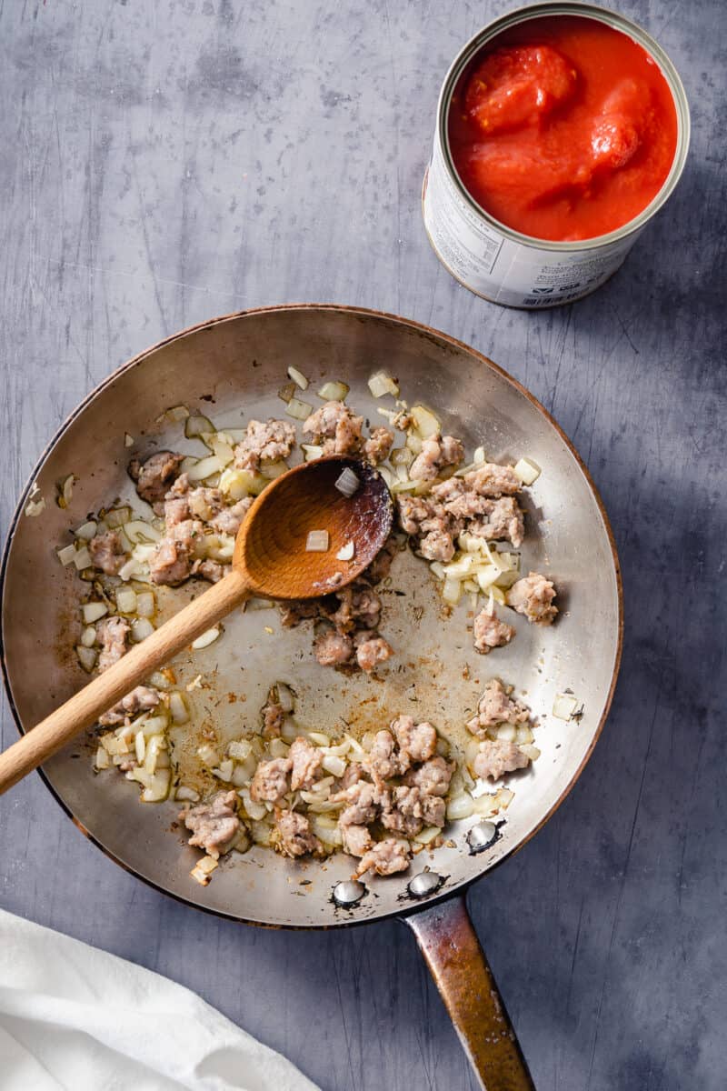 Diced onions, crumbled Italian sausage, and sliced garlic in a skillet with a can of tomatoes nearby