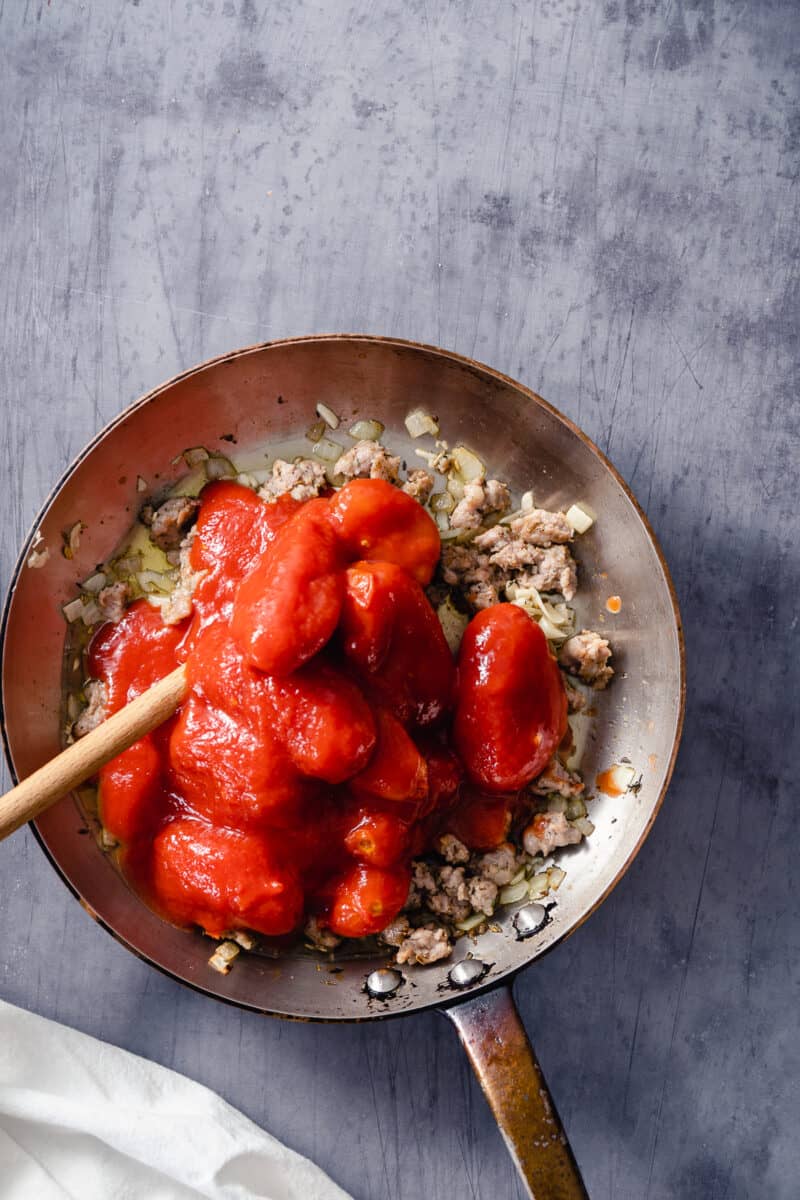 Whole peeled tomatoes added to a skillet with onions, garlic, and sausage