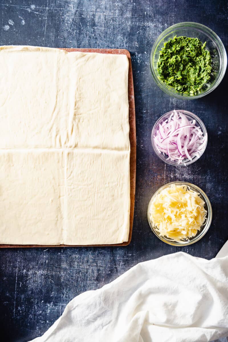 A sheet of puff pastry, green chutney, red onions, and cheddar cheese in bowls