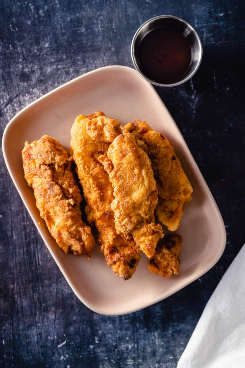 Crispy chicken tenders on a pink plate with a container of dipping sauce next to it