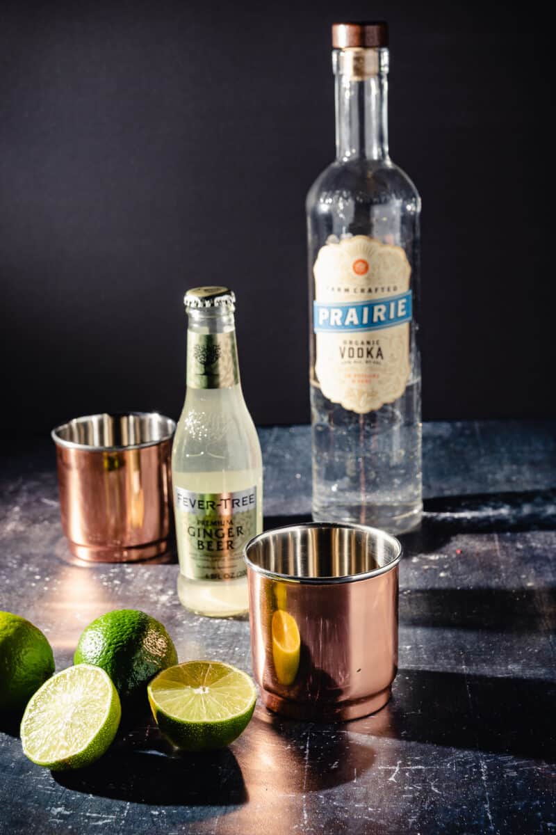 Limes sliced in half next to a copper tumbler with bottles of ginger beer and vodka in the frame