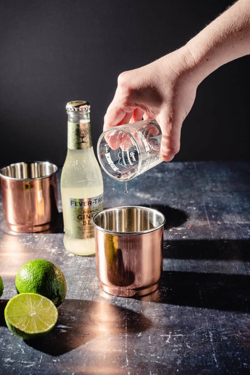 A drop of vodka clings to a measuring glass above a copper cup