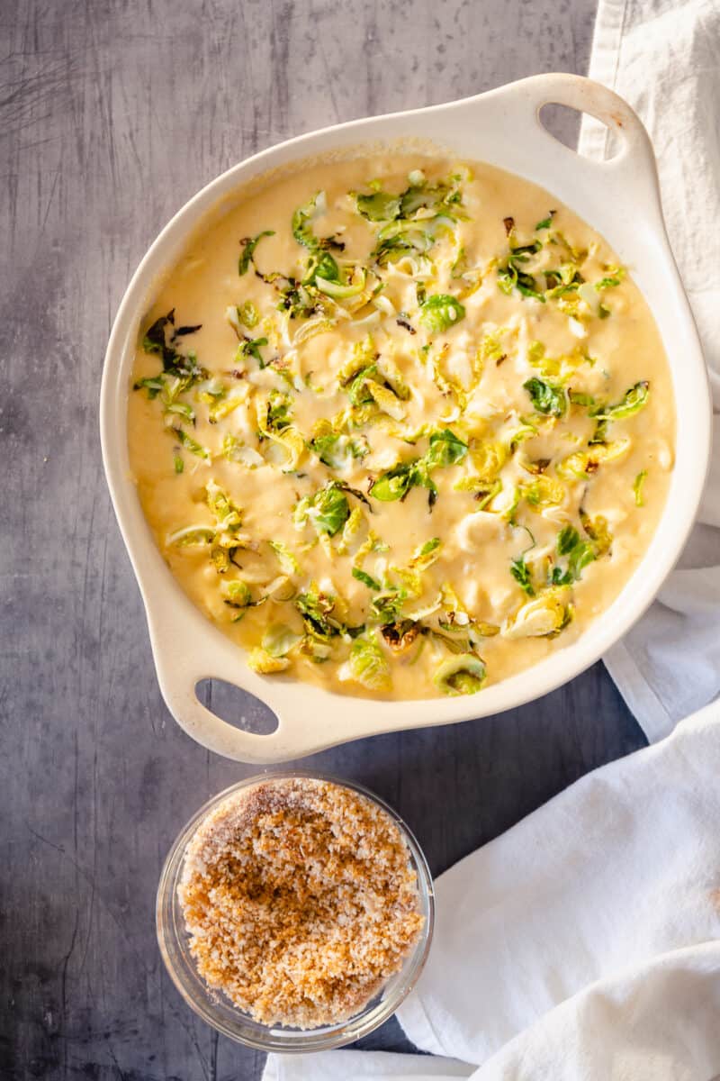 Sliced, roasted Brussels sprouts added to pasta and cheese sauce in a casserole dish with breadcrumb mixture on the side