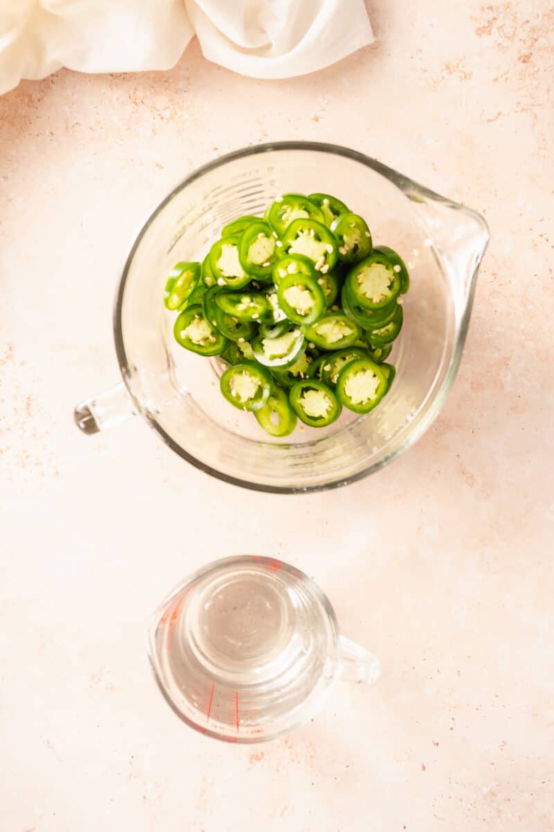 Sliced jalapeños in a bowl with a measuring cup with white vinegar alongside