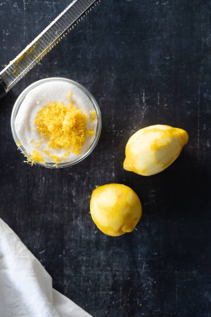 Lemon zest grated into a bowl of sugar with zested lemons alongside
